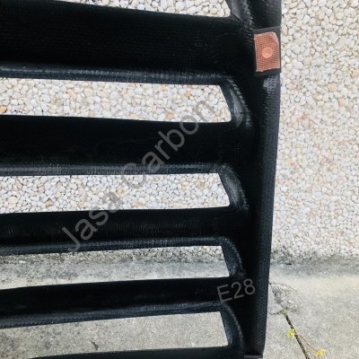 Estrutura preta tipo grade com barras horizontais inclinadas em material carbono, contra parede de seixos brancos.