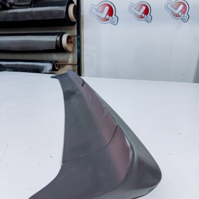 Spoiler automóvel em carbono preto sobre superfície branca com rolos de tecido de carbono ao fundo e texto Jasarcarbon.com na parede.