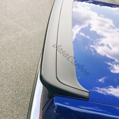 Spoiler preto com texto JasaCarbon numa parte traseira azul de carro, junto a farolim laranja e carros estacionados.