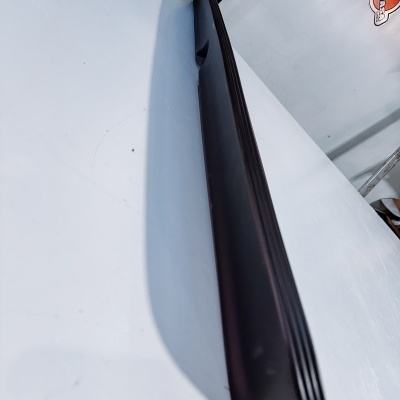 Barra preta longa, possivelmente soleira de porta de carro, em superfície branca com adesivos na parede ao fundo