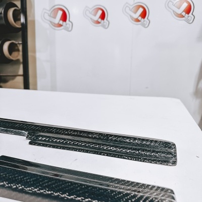 Produtos de fibra de carbono preta com textura sobre mesa branca, painel com texto e logótipos ao fundo.