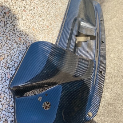 Peça automóvel em fibra de carbono com padrão quadriculado sobre chão de seixos