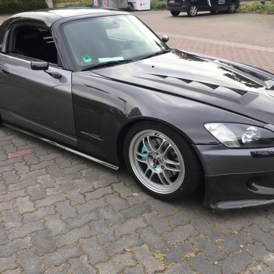 Carro desportivo cinza metálico com capota preta estacionado numa superfície de pavê