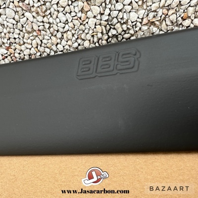 Painel preto com logo BBS em relevo sobre base de madeira clara e cascalho ao fundo