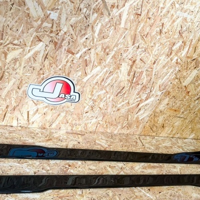 Dois protetores laterais pretos para carro sobre superfície de madeira OSB com autocolantes Japs