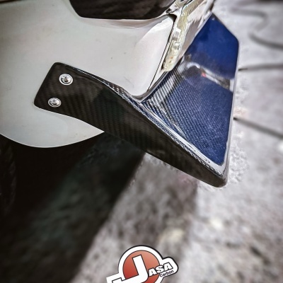 Peça automóvel em fibra de carbono preta fixada num veículo branco com texto promocional.