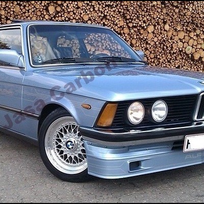 Carro clássico BMW azul claro com jantes prateadas e fundo de madeira empilhada.