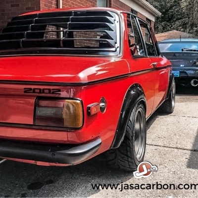 Carro vermelho BMW 2002 estacionado em entrada de garagem com outra viatura ao fundo
