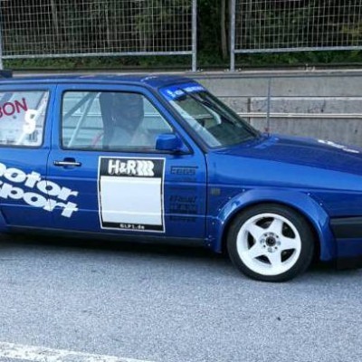 Carro de corrida azul com autocolantes e rodas brancas estacionado numa estrada