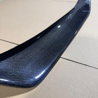 Spoiler de automóvel em fibra de carbono preta com textura visível, sobre cartão castanho