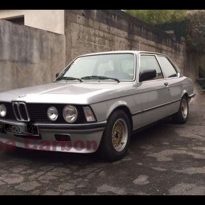 Carro BMW clássico prata estacionado junto a muro de pedra e edifício
