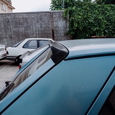 Antena preta em carro cinza estacionado, com outros carros e parede ao fundo