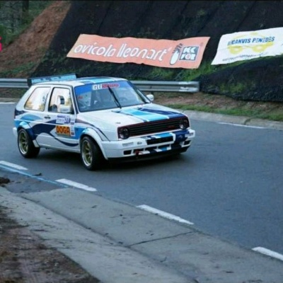 Carro de rali branco com detalhes em azul e vermelho numa curva de estrada asfaltada com cartazes publicitários ao fundo.
