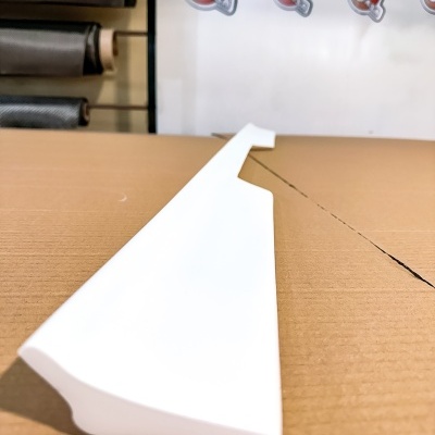 Peça branca triangular sobre papelão com rolos ao fundo e placa jasacarbon.com