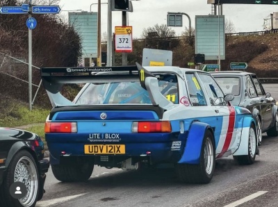 Carro de corrida azul claro com faixa vermelha e branca, asa traseira preta e placa amarela em estrada