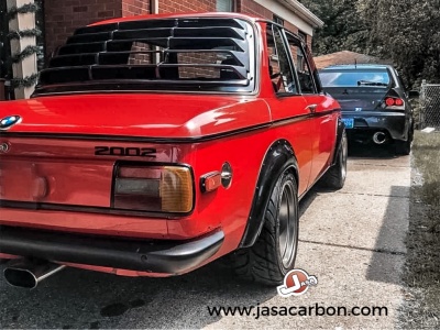 Carro vermelho BMW 2002 estacionado em entrada de garagem com outra viatura ao fundo