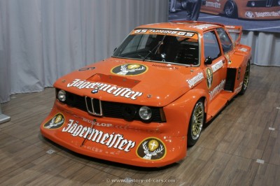 Carro de corrida BMW laranja com logótipo Jägermeister exposto em interior.