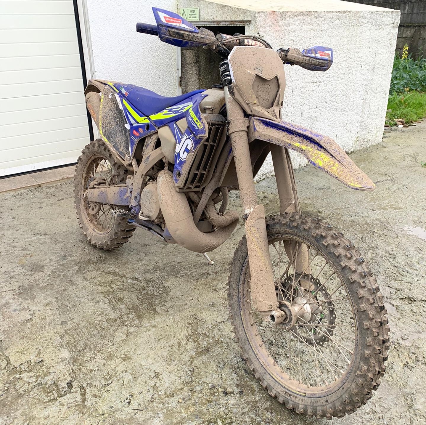 Motocross azul e amarelo coberta de lama com pneus de cravos em chão de cimento junto a parede branca