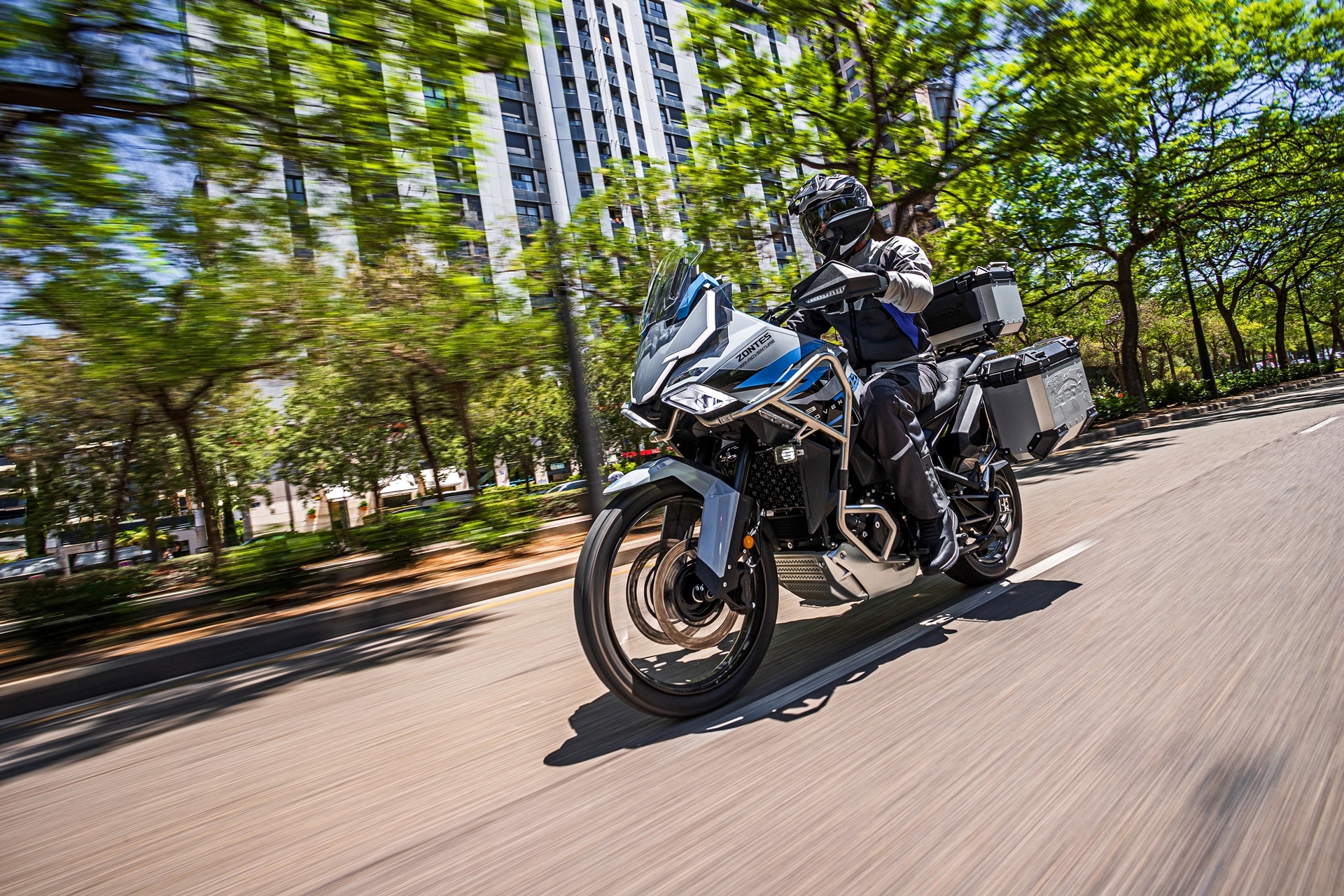 Motocicleta de turismo cinza com malas rígidas e piloto em estrada urbana