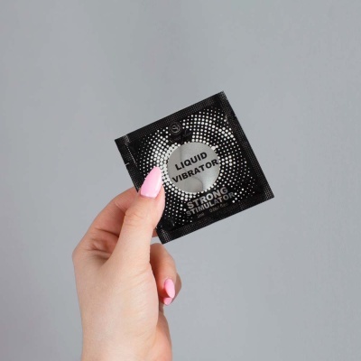 Pacote quadrado preto com texto Liquid Vibrator, segurado por mãos femininas com unhas pintadas