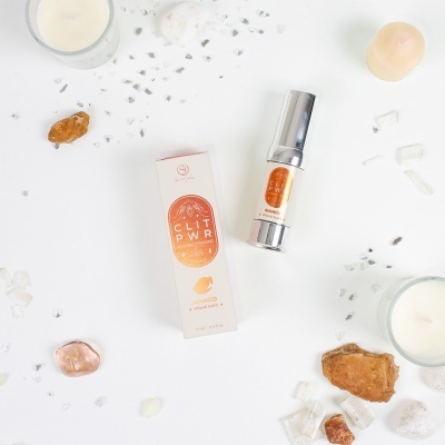 Produto cosmético CLIT POWR MANGO com pedras e velas brancas em fundo branco