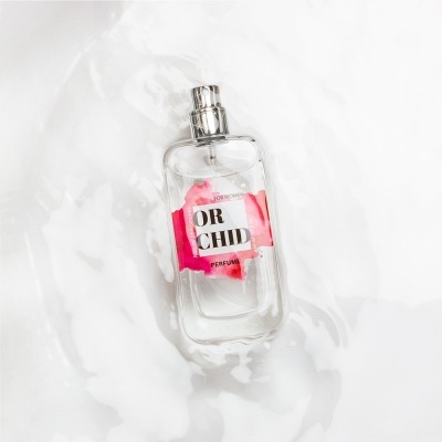 Frasco de perfume transparente com rótulo rosa e texto em fundo branco com espuma em torno.