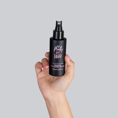 Frasco preto de spray de óleo seco com rótulo em rosa e branco, segurado por mão com unhas rosa claro