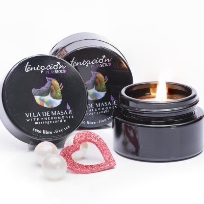 Frasco preto com vela de massagem acesa, tampa com rótulo preto, pérolas brancas e coração vermelho sobre fundo branco