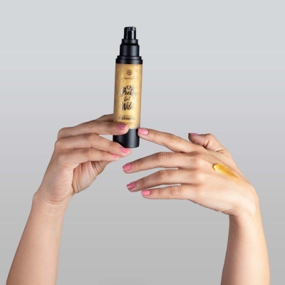 Frasco de produto dourado com bomba preta segurado por mãos femininas com amostra no dorso, fundo cinza claro.