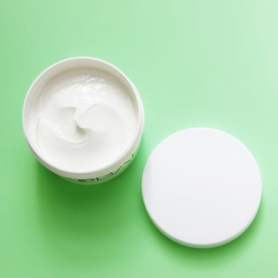 Pote aberto de creme branco com tampa branca ao lado sobre fundo verde claro