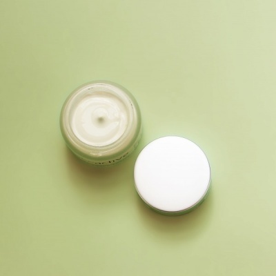 Frasco aberto de creme branco com tampa ao lado sobre fundo verde claro