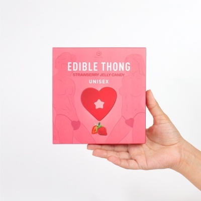 Embalagem de papel rosa com texto 'EDIBLE THONG' e ilustração de morangos, segurada por uma mão.