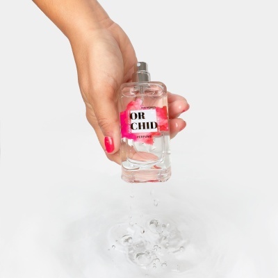 Frasco de perfume transparente com rótulo colorido com a palavra OR CHID, segurado por mão com unhas vermelhas derramando líquido