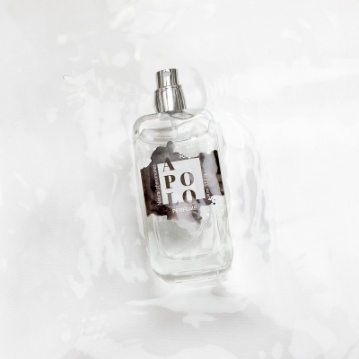 Frasco de perfume transparente com rótulo 'APOLO' e tampa prateada sobre fundo branco texturizado