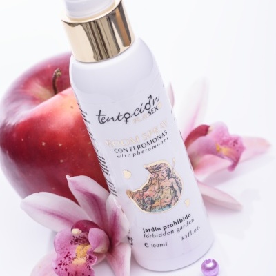 Frasco de spray branco com rótulo decorativo ao lado de maçã, flores rosa e pérolas lilases.