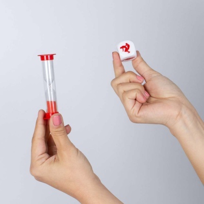 Temporizador de areia com bases vermelhas e peça branca com símbolo vermelho, segurados por mãos.