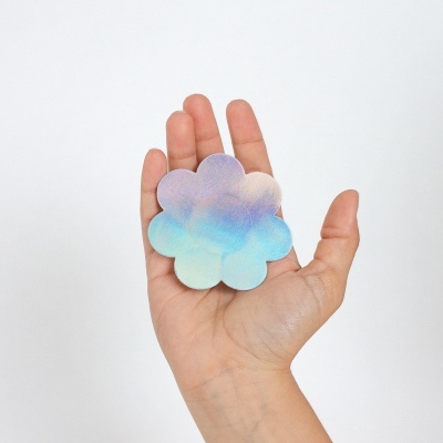 Adesivo em forma de flor com cores iridescentes segurado por uma mão sobre fundo branco