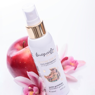 Frasco branco de spray com texto em espanhol, maçã vermelha e flores cor-de-rosa ao lado.