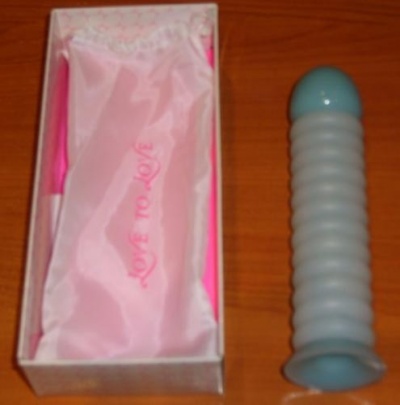 Objeto cilíndrico cinza claro com base azulada ao lado de uma caixa com saco rosa satinada com inscrição 'LOVE TO LOVE'