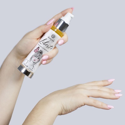 Frasco de creme com rótulo ilustrado sendo segurado por mão com unhas cor rosa, aplicado na outra mão