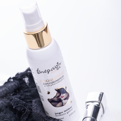 Frasco branco de spray corporal com tampa dourada e texto em espanhol, sobre renda preta, com tampa prateada ao lado