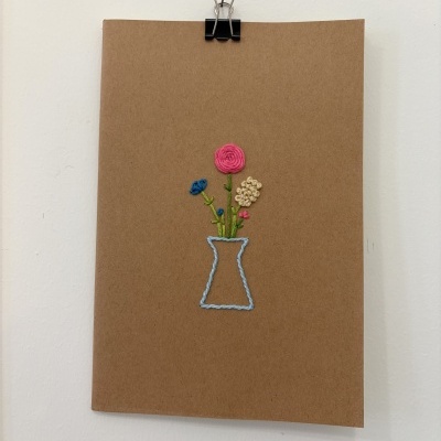Cartão castanho com bordado de flores coloridas em vaso azul claro, pendurado por clipe preto
