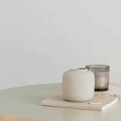 Mesa redonda branca com dois vasos decorativos sobre um livro, ao lado de cadeira com assento de palha.