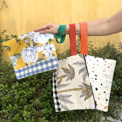 Três sacolas de tecido com padrões florais e abstratos seguradas por uma mão contra fundo amarelo e vegetação