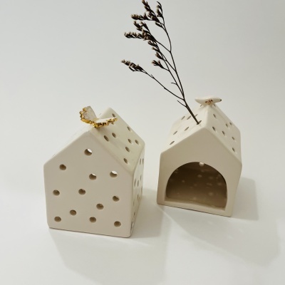 Dois adornos decorativos em forma de casinha em cerâmica branca com pequenos furos e um anel dourado e ramo seco.