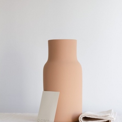Vaso de cerâmica bege, cartão com texto 'o cactuu' e pano branco sobre mesa branca.