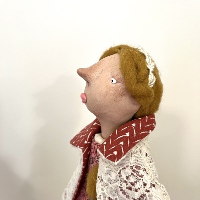 Boneca artesanal com rosto tecido, cabelo lã castanho, capa de renda branca e casaco vermelho com padrão branco.