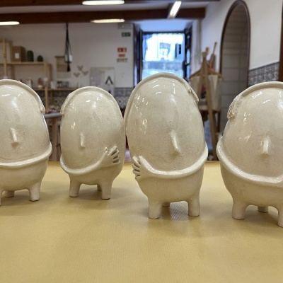 Quatro estatuetas em cerâmica bege com rostos simples, sobre superfície amarela em ambiente interior