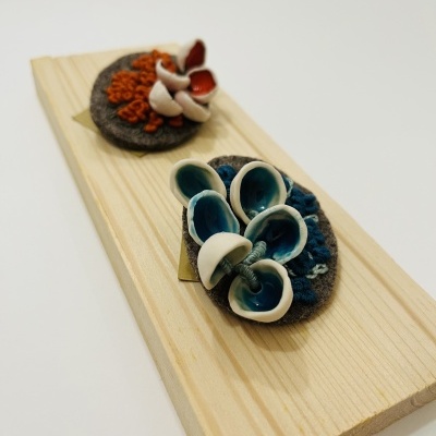 Duas peças de bijuteria artesanais em base de tecido cinzento com elementos cerâmicos coloridos sobre tábua de madeira.