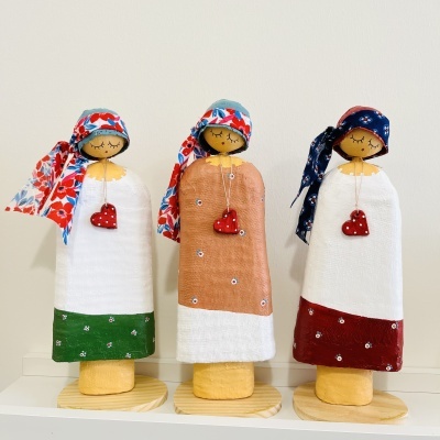 Três bonecas decorativas de madeira e tecido com lenços coloridos e coração vermelho no pescoço em prateleira branca